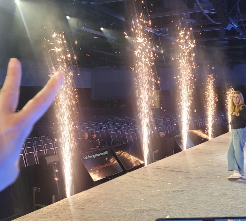 Stage-level view of cold spark fountains firing, with a DJ's hand raised in a two-finger peace sign in the foreground