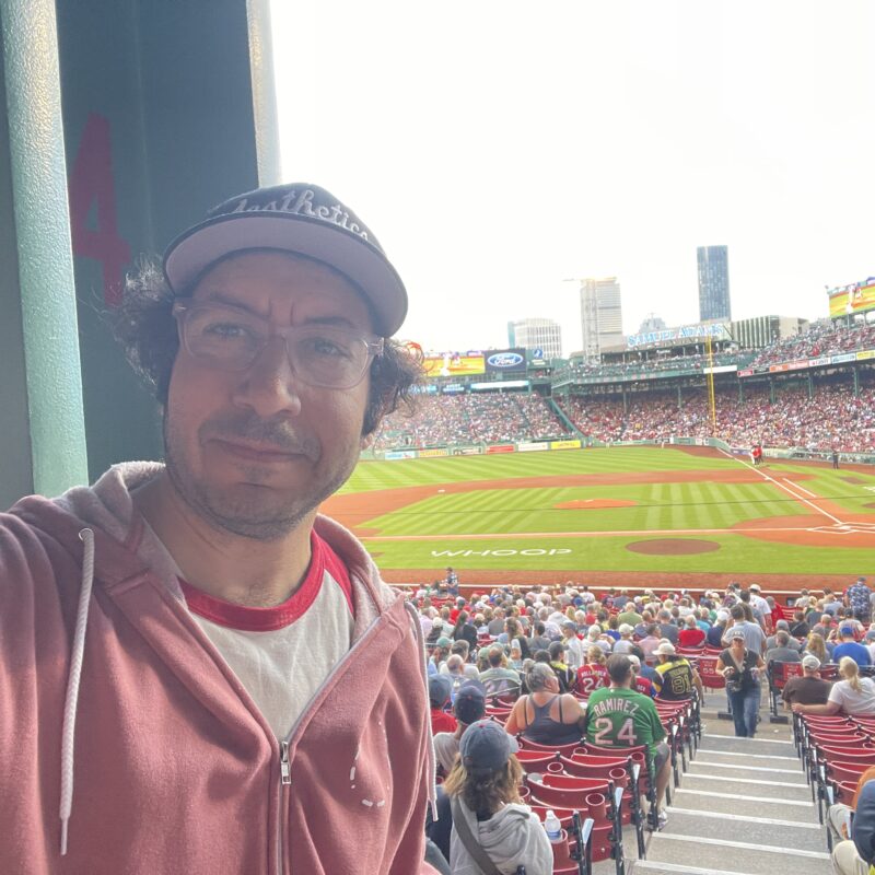 Sean Gannet takes a selfie beside a wide green support column at Fenway Park, with the infield and a packed grandstand visible past it
