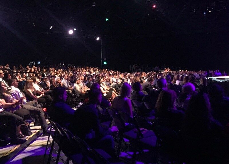 A speaker on a long runway stage addressing a packed audience in a darkened venue, seen from among the seats